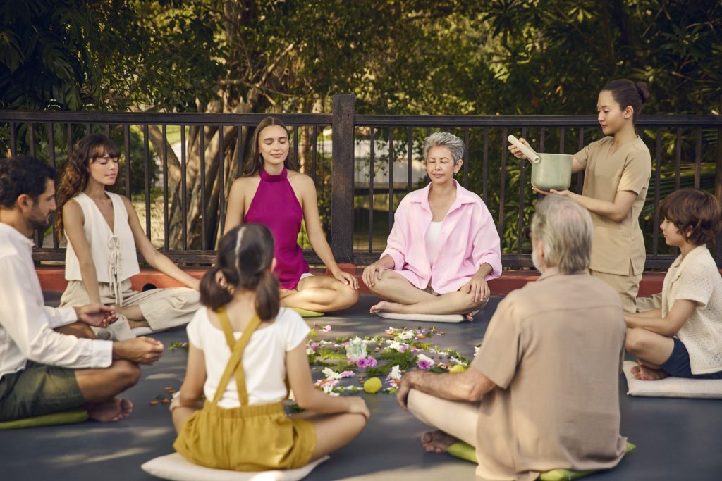 A wellness group meditation session at RXV Wellness Village, featuring guests of all ages sitting in a circle with a floral mandala, guided by a therapist in a serene garden setting.