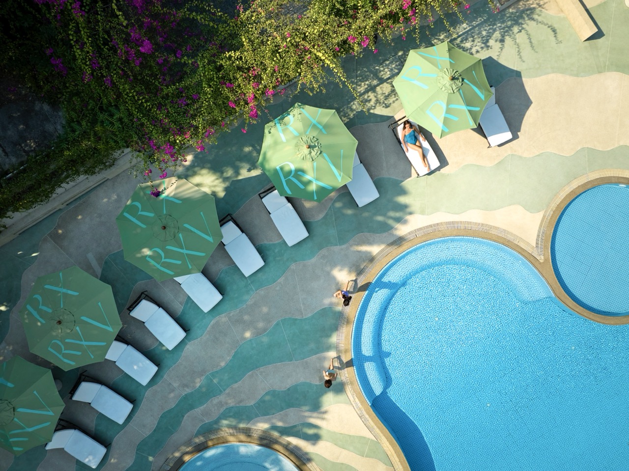 Aerial view of RXV Wellness Village Sampran swimming pool with turquoise umbrellas and sun loungers surrounded by lush greenery.