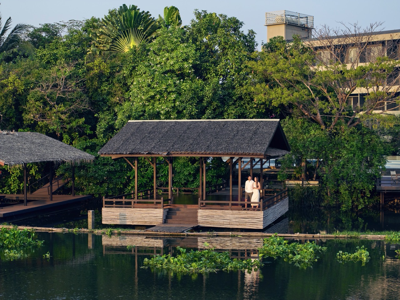 Couple standing on a riverside pavilion surrounded by lush tropical greenery at RXV Wellness Village Sampran.